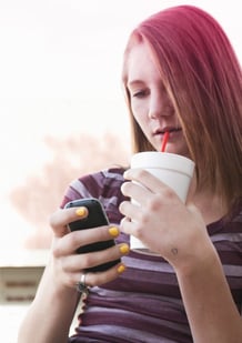 Kuva tytöstä juomassa pillillä mukista ja käyttämässä kännykkäänsä.
